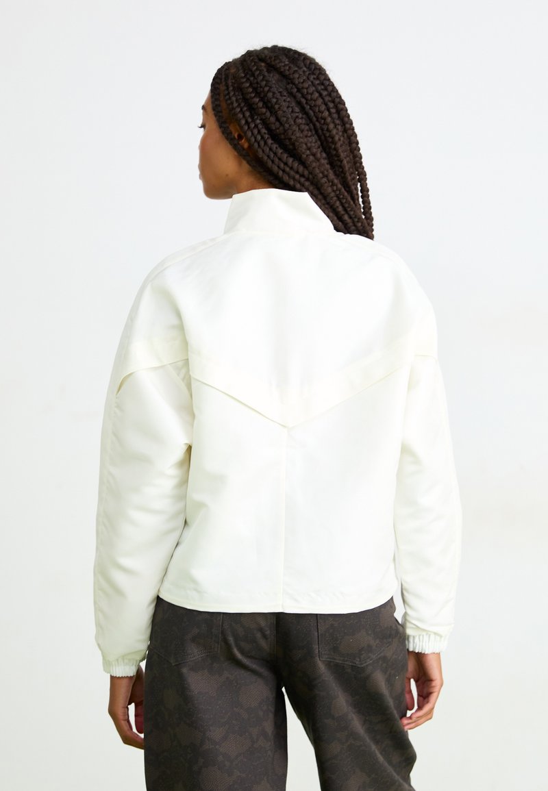 Woman with long braids wearing a white jacket and dark patterned pants, facing away against a plain white background.