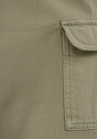 Tuvplāna gaiši brūna auduma tuvplāns, kurā redzama sīkākas šuvju detaļas un flapa kabata ar pastiprinātiem malām labajā pusē.