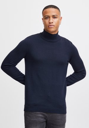 Navyblauwe coltrui van een zachte brei stof, met lange mouwen en een ribgebreide zoom, gedragen over een donkere spijkerbroek.
