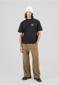 Czarny bawełniany t-shirt z krótkimi rękawami i logo z przodu. Połączony z szerokimi, beżowymi spodniami i czarnymi butami. Model nosi kremową wełnianą czapkę.