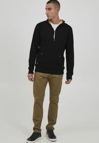 Felpa nera con zip e colletto interno giallo, due tasche frontali, abbinata a pantaloni beige e sneakers nere. Look casual.