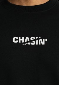 Juodas medvilnės džemperis su baltu užrašu "CHASIN'", kuriame yra įstrižas brūkšnis per "CHAS" kaip grafinis akcentas. Apvalus kaklas ir įtempiančios medžiagos.
