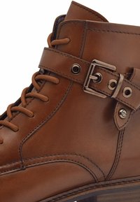 Botte de cheville en cuir marron avec surface texturée, boucle en métal et œillets décoratifs, présentant un design classique à lacets et une semelle robuste.