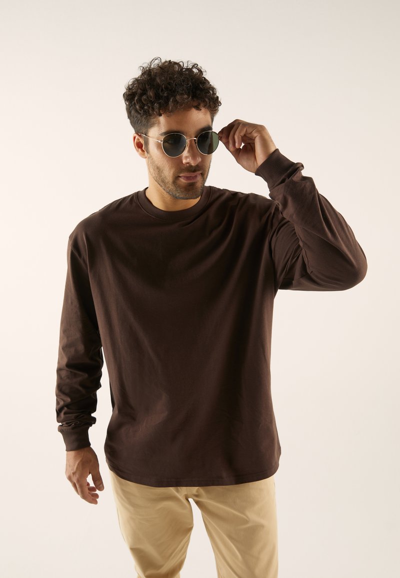 T-shirt marron à manches longues avec un col rond, texture douce, associé à un pantalon beige clair et des lunettes de soleil rondes. Design simple et décontracté.