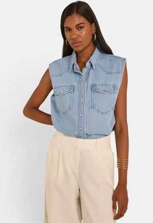 Vrouw draagt een mouwloos lichtblauw denimshirt met knoopsluitingen op de borstzakken en beige hoge taille broeken, poseert met één gebogen arm.