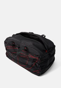 Sac de sport noir avec des coutures d'accent rouge, fabriqué dans un matériau durable, avec des poignées doubles, une fermeture zippée et des poches latérales.