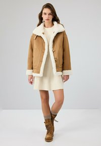 Bruine shearling jas met witte bontkraag, knoopsluiting en twee voorzakken. Model draagt een crème ribbeljurk en bruine veterschoenen.