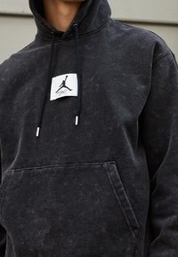 Sudadera negra de tejido texturizado con un bolsillo canguro, que presenta una etiqueta blanca con un logotipo negro y cordones con puntas plateadas.