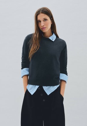 Jeune femme aux longs cheveux bruns portant un pull noir par-dessus une chemise rayée bleue et un pantalon noir, les mains dans les poches sur un fond uni.
