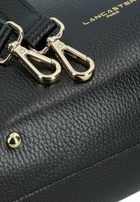 Borsa a mano in pelle nera con hardware dorato, doppio moschettone e superficie texturizzata. Include dettaglio con il marchio: "Lancaster Paris."