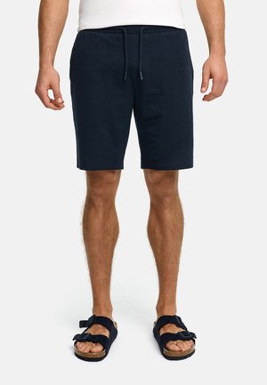 Mężczyzna w granatowych szortach z troczkami i czarnych sandałach z odkrytymi palcami, stojący na tle jednolitego tła.