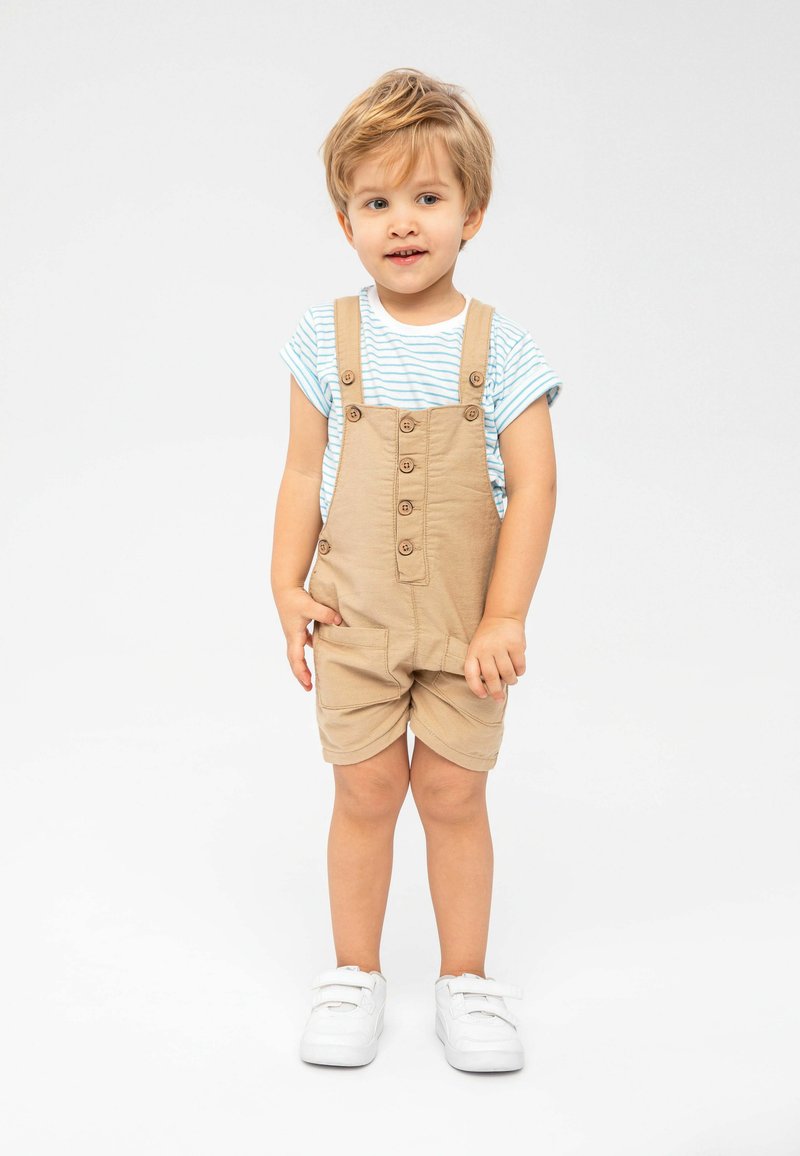 Kohvased overalls, millel on nupud ja eesmine tasku, kantud sinise ja valge triibulise särk üle. Valged spordijalatsid täiendavad komplekti.