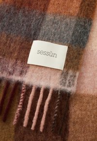Wollen deken met een geruit patroon in tinten bruin, roze en grijs. Heeft franjes aan de randen en een wit stoffen label met "sessùn" erop gedrukt.