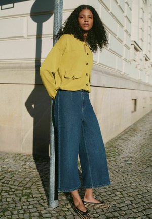 Młoda kobieta o kręconych włosach, ubrana w żółtą koszulę na guziki, szerokie niebieskie dżinsy i wzorzyste baleriny, stoi przy słupie na ulicy wyłożonej kamienną kostką.