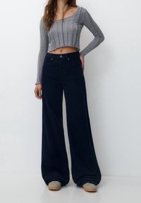 Un haut long à manches longues côtelé gris, coupé court, associé à un pantalon large bleu marine taille haute. Les matériaux et textures ne sont pas spécifiés. Le pantalon est doté d'une fermeture à bouton.