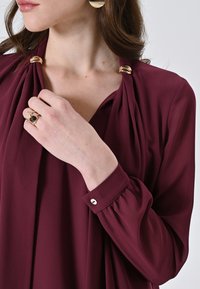 Blusa bordeaux con tessuto drappeggiato, dettagli in metallo dorato sulle spalle e bottone sui polsini; texture liscia e design fluido.