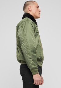 Alyvuogių žalia bombonešio striukė su juodo dirbtinio kailio apykakle, užtrauktuku priekyje, ribotais rankogaliais ir apačia bei šoninėmis kišenėmis su lopais.