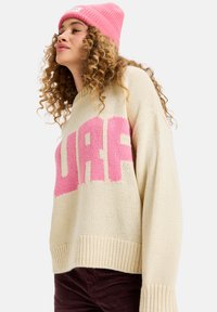 Crèmekleurige gebreide trui met een oversized pasvorm, voorzien van grote roze "SURF" letters. Ribgebreide manchetten en onderrand, gedragen met een roze beanie.