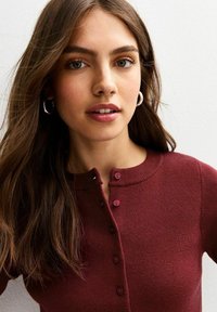 Bordeaux geribbelde cardigan met een ronde halslijn, kleine knopen en een aansluitend ontwerp. Heeft een gladde textuur en subtiele details.