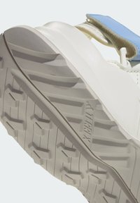 Λευκή σόλα αθλητικού παπουτσιού με υφασμένο ανάγλυφο σχέδιο πέλματος, με μπλε λουράκι και λείο ανώτερο υλικό. Το όνομα της μάρκας «TERREX» φαίνεται στο πλάι.