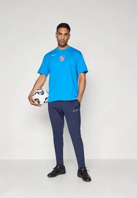 Camiseta atlética azul com o logótipo da equipa, calças de treino azul-escuro e sapatos pretos; segurando uma bola de futebol estampada. Design de roupa desportiva casual.