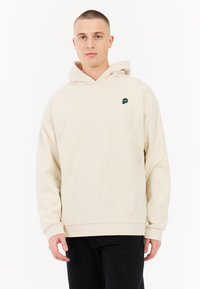 Beige hoodie met een trekkoord, kangoeroezak en een groen logo op de linkerkant van de borst. Zachte textuur, relaxte pasvorm, ribgebreide manchetten en zoom.