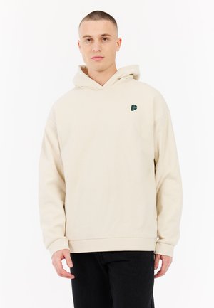Beige hoodie met een trekkoord, kangoeroezak en een groen logo op de linkerkant van de borst. Zachte textuur, relaxte pasvorm, ribgebreide manchetten en zoom.