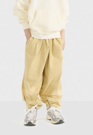 Helle, gelbe Baumwoll-Jogginghosen mit einem elastischen Bund, seitlichen Taschen und einem lockeren Schnitt, kombiniert mit grauen und cremfarbenen Sneakern.