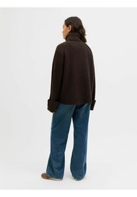 Maglione oversize marrone scuro con colletto alto e polsini a coste, abbinato a jeans in denim blu a gamba larga e scarpe marroni.