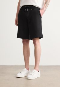 Černé bavlněné šortky s elastickým pasem a šňůrkou. Disponují bočními kapsami a čistým, minimalistickým designem. Nosič má na sobě bílé tenisky.