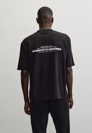 Mann mit kurzen Haaren und Ohrringen, der ein schwarzes T-Shirt mit weißem Text "Moments to Memories" auf dem Rücken vor einem einfarbigen hellen Hintergrund trägt.