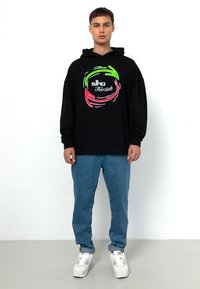 Schwarzer Hoodie mit buntem Grafiklogo, Kängurutasche, gerippten Bündchen, kombiniert mit hellblauen Jeans und weißen Sneakers. Schlichter, lässiger Look.