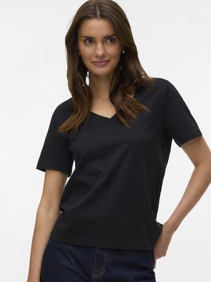 Sieviete ar gariem brūniem matiem, valkājot melnu V veida kakla īsroku krekliņu un tumši zilas džinsas, pozē ar vienu roku uz gurniem baltā fona.