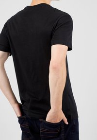 Camiseta de algodón negra con cuello redondo, mangas cortas y textura suave, vista desde la espalda con las manos en los bolsillos de unos jeans oscuros.