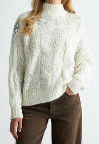 Žena oblečená v bílém pleteném svetru s textura a flitrovými nášivkami na rameni a hrudi, doplněná o tmavě hnědé kalhoty, ruce v uvolněné poloze.