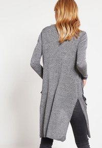 Grå långärmad topp med en texturerad stickning, med sidofickor och en avslappnad passform, ihop med mörka jeans, sedd bakifrån.