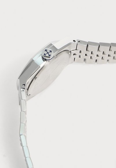 Montre en métal argenté avec un bracelet à maillons et une couronne présentant un logo à quatre flèches sur un fond clair uni.