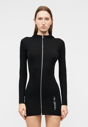 Donna in piedi di fronte che indossa un vestito nero aderente a maniche lunghe con una cerniera frontale e testo bianco vicino all'orlo.