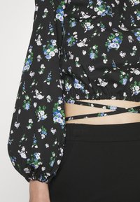 Crop top noir à imprimé floral avec manches longues bouffantes et liens croisés à la taille, porté avec un pantalon noir taille haute.