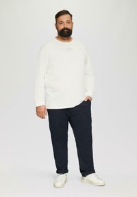 Weißes Langarmhemd mit kleinem Logo, kombiniert mit dunkelblauen Hosen und weißen Sneakers. Einfaches Design, Baumwollmaterial, lockere Passform.