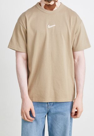Henkilö, joka käyttää beigeä Nike-t-paitaa, jossa on valkoinen Swoosh-logo, ja vaaleansinisiä farkkuja, seisoo yksinkertaista vaaleaa taustaa vasten.