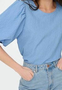 Top azzurro chiaro con texture, maniche corte a sbuffo, scollatura rotonda, abbinato a pantaloni corti in denim a vita alta con chiusura a bottone e tasche.