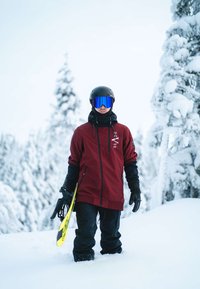 Red snowboard jacket, black pants, and gloves worn by a person holding a yellow snowboard. Black helmet and blue mirrored goggles. Snowy background.