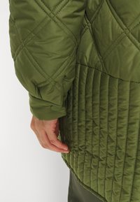 Chaqueta acolchada verde tipo plumas con costuras en forma de diamante y verticales. Presenta un dobladillo redondeado y detalles de puño abotonado en la manga.