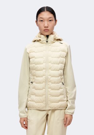 Frau mit glatten dunklen Haaren trägt eine hellbeige gesteppte Jacke mit Reißverschlusstaschen und passende Hosen, blickt nach vorne vor einfarbigem Hintergrund.