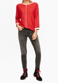 Pull en tricot rouge avec des accents blancs, associé à un jean gris slim et des bottes noires avec des lacets rouges et des œillets métalliques.