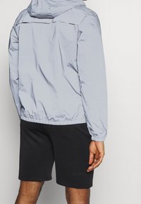 Chaqueta con capucha de color azul claro con puños y dobladillo elásticos, llevada sobre pantalones cortos negros que presentan un sutil detalle estampado. Tejido suave y ligero.