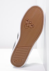 La suola della sneaker presenta una suola in gomma marrone con motivo a spina di pesce, una intersuola bianca e un logo impresso nell'area del tallone.