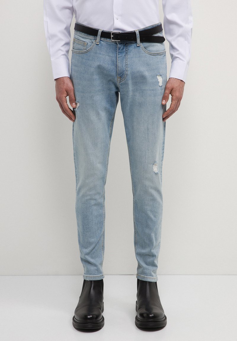 Jeans in denim azzurro chiaro con vestibilità slim, caratterizzati da dettagli strappati, cinque tasche e una cintura nera indossati con una camicia bianca con bottoni.