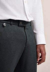 Dunkelgraue, strukturierte Hose mit Slim-Fit, ausgestattet mit zwei Metallknöpfen und einem glatten Bund, kombiniert mit einem weißen Langarmhemd.